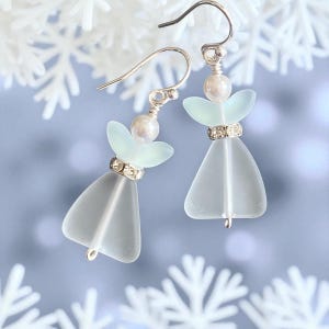 May include: A pair of angel-shaped earrings. Each earring features a frosted glass body, a pearl bead, and a halo of pale green glass. The earrings have silver-colored hooks and a band of rhinestones around the waist of the angel.