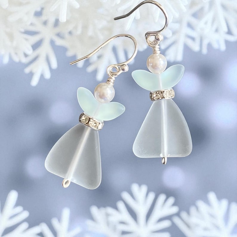 May include: A pair of angel-shaped earrings. Each earring features a frosted glass body, a pearl bead, and a halo of pale green glass. The earrings have silver-colored hooks and a band of rhinestones around the waist of the angel.