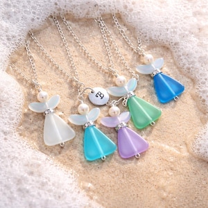 Puede incluir: Conjunto de collares con colgantes de &aacute;ngel en varios colores: blanco, azul, turquesa, morado y verde. Cada collar tiene una perla como cabeza, detalles de pedrer&iacute;a y una cadena plateada. Los colgantes est&aacute;n sobre una superficie arenosa.