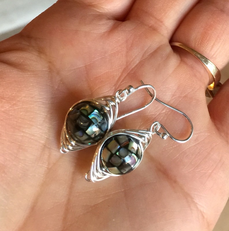 Op de afbeelding: Een paar zilveren oorbellen met een zwarte en iriserende abalone-schelpparel die is gewikkeld in zilverdraad.