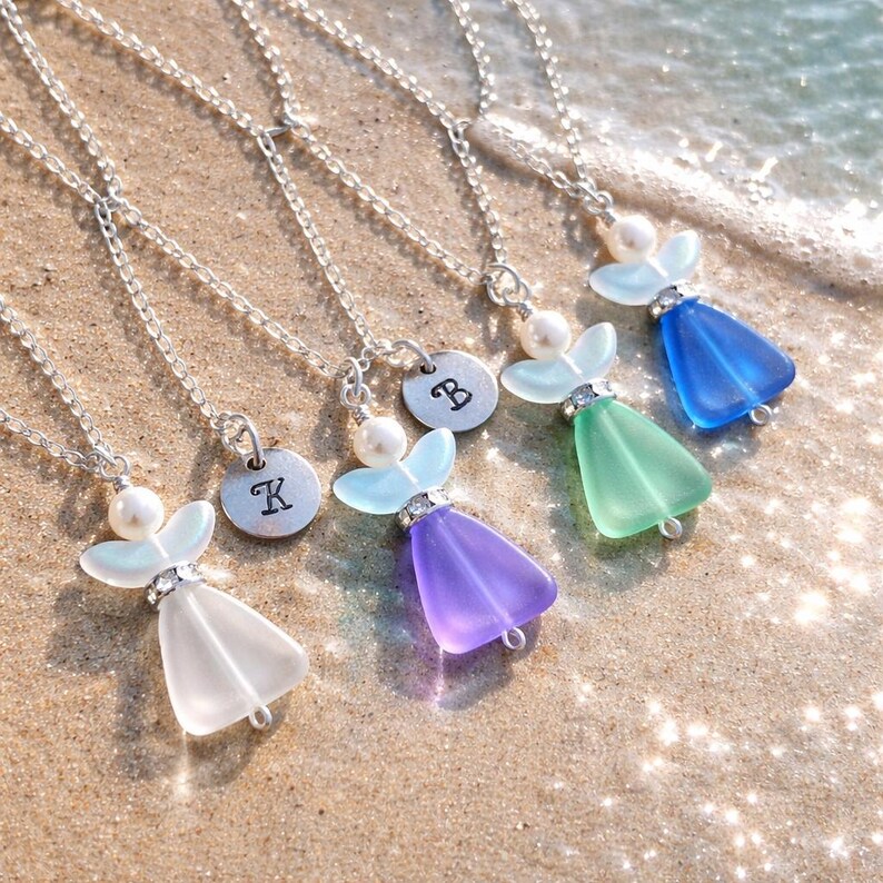 Puede incluir: Conjunto de collares con colgantes de alas de &aacute;ngel y cadenas plateadas. Cada uno presenta un vestido de cristal de color diferente y una cabeza de perla. Algunos tienen un peque&ntilde;o dije redondo con una letra. Sobre arena.