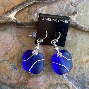 May include: A pair of sterling silver earrings featuring vibrant blue sea glass. Each piece of sea glass is wrapped with silver wire and suspended from a silver hook. The earrings are displayed on a black card that reads "STERLING SILVER".