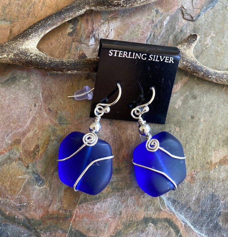 May include: A pair of sterling silver earrings featuring vibrant blue sea glass. Each piece of sea glass is wrapped with silver wire and suspended from a silver hook. The earrings are displayed on a black card that reads "STERLING SILVER".