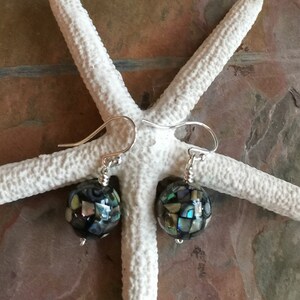 Op de afbeelding: Een paar zilveren oorhangers met iriserende abalone schelp kralen. De kralen zijn rond en hebben een moza&iuml;ekpatroon.