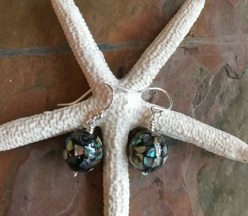 Op de afbeelding: Een paar zilveren oorhangers met iriserende abalone schelp kralen. De kralen zijn rond en hebben een moza&iuml;ekpatroon.