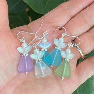 May include: Five pairs of angel-shaped earrings in various pastel colors: lavender, white, turquoise, light blue, and green. Each earring features a pearl bead, a rhinestone-studded band, and silver-tone ear wires. The wings are iridescent.