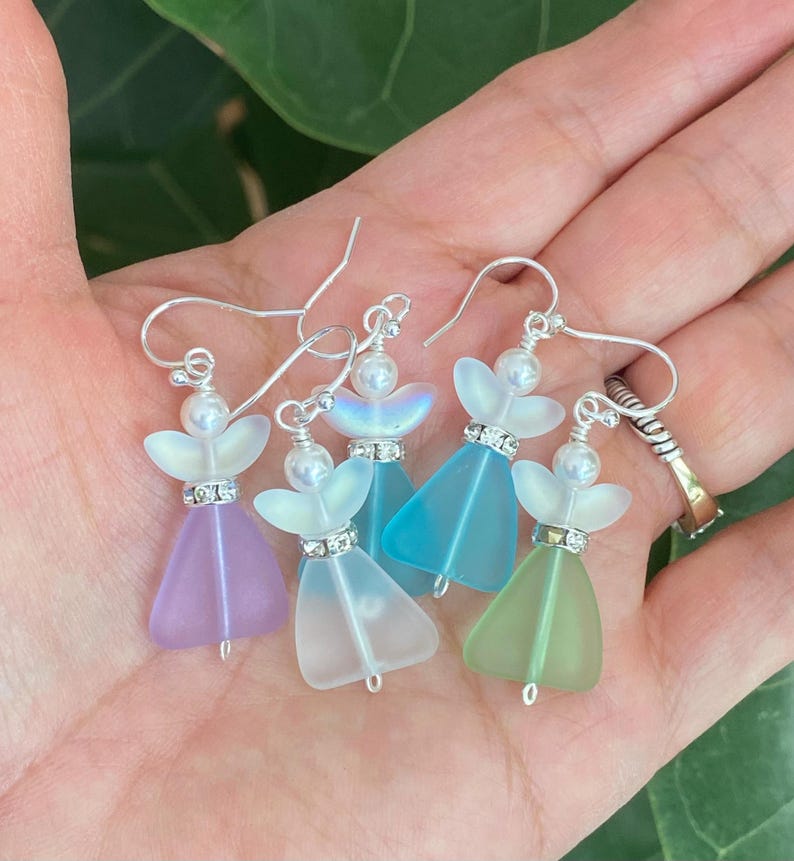 May include: Five pairs of angel-shaped earrings in various pastel colors: lavender, white, turquoise, light blue, and green. Each earring features a pearl bead, a rhinestone-studded band, and silver-tone ear wires. The wings are iridescent.