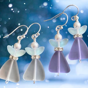 May include: Four pairs of angel-shaped earrings with silver hooks. Each earring features a pearlescent bead, a pair of light blue wings, and a crystal-studded band above a translucent, bell-shaped body in shades of gray and purple.