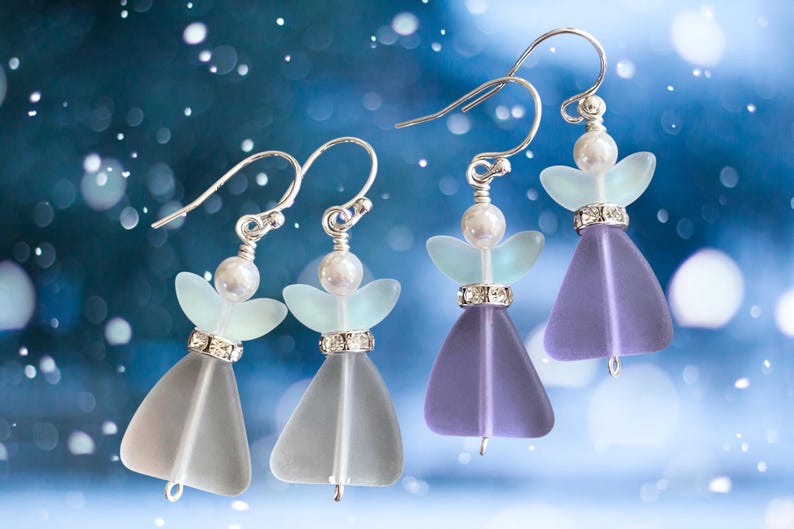 May include: Four pairs of angel-shaped earrings with silver hooks. Each earring features a pearlescent bead, a pair of light blue wings, and a crystal-studded band above a translucent, bell-shaped body in shades of gray and purple.