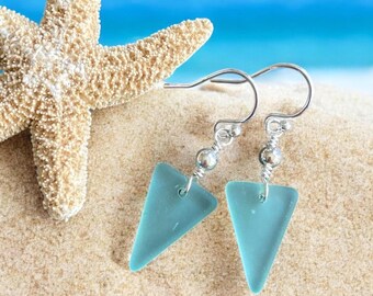 May include: A pair of turquoise triangle earrings with silver-colored hooks and wire detailing. The earrings are displayed on a sandy surface with a starfish in the background, evoking a beach theme.