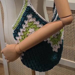 May include: A handmade crochet bag in shades of teal, white, and green. The bag features a granny square design and a braided strap. The bag is displayed on a mannequin.