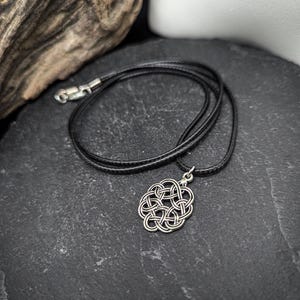 Può includere: Una collana con pendente a nodo celtico color argento e cordino nero. Il pendente presenta un intricato design intrecciato. La collana è esposta su una superficie scura e strutturata, con legno alla deriva sullo sfondo.