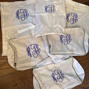 May include: A collection of white travel bags and packing cubes with a blue monogram. The bags vary in size and style, including mesh and solid fabric options. The monogram is embroidered on each bag, adding a personalized touch to the travel accessories.
