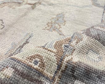 May include: Close-up of a light-colored area rug with a detailed pattern. The rug features a mix of beige, gray, and muted blue tones. The design includes floral and geometric elements, creating a textured, vintage aesthetic.