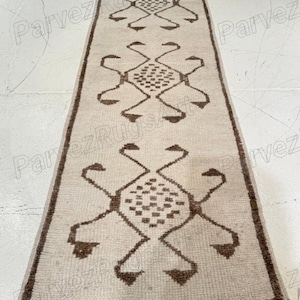 May include: A rectangular beige runner rug with a repeating geometric pattern in brown. The rug has a border and the pattern includes diamond shapes and stylized elements. The rug is designed for a hallway or entryway.