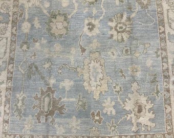 May include: A light blue and beige patterned rug with floral and geometric designs. The rug has a distressed look with a border. White sandals with gold accents are visible.