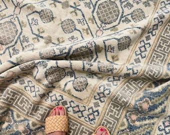May include: Beige and blue patterned rug with a geometric border. The rug features a repeating floral design in shades of gray, blue, and pink. Woven leather sandals are visible on the rug.