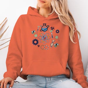 May include: Orange hoodie featuring a science-themed graphic. The graphic includes colorful illustrations of atoms, beakers, DNA strands, and other scientific symbols. The hoodie has a drawstring hood and a front pocket.
