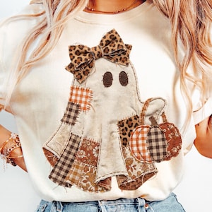Puede incluir: Camiseta color crema con un diseño de fantasma de patchwork y un lazo con estampado de leopardo. El fantasma sostiene una bolsa con forma de calabaza. El patchwork incluye patrones marrones, naranjas y crema, ideal para Halloween.