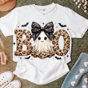 May include: A white short-sleeve t-shirt featuring a graphic of a ghost wearing a large black lace bow. The ghost is positioned between the letters B and O, which are filled with a brown leopard print pattern. Small black bats and stars surround the central design.