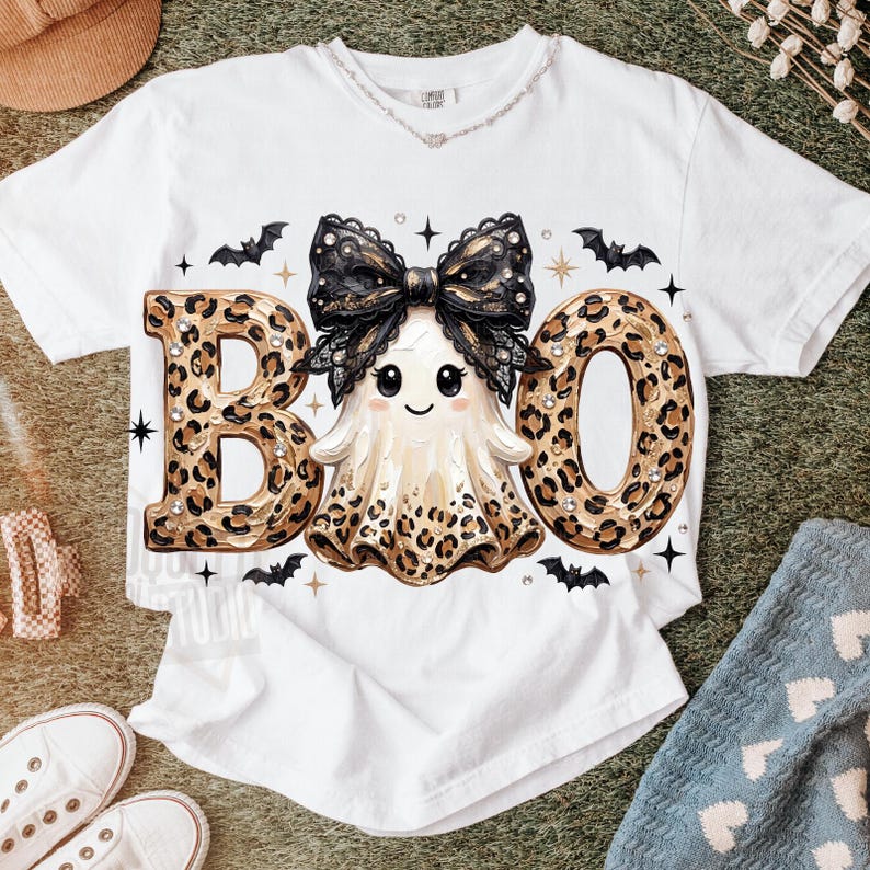May include: A white short-sleeve t-shirt featuring a graphic of a ghost wearing a large black lace bow. The ghost is positioned between the letters B and O, which are filled with a brown leopard print pattern. Small black bats and stars surround the central design.