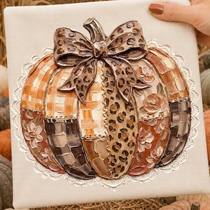 Pode incluir: Uma abóbora decorativa com um laço, apresentando um design de patchwork em tons de laranja, castanho e creme. O laço tem um padrão de leopardo. A abóbora é adornada com detalhes florais e uma borda de renda.