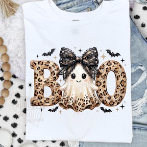 May include: A white t-shirt featuring a graphic print of the word BOO. The letters B and O are filled with a brown and black leopard print pattern. A central ghost character wears a large black lace bow and has a leopard print skirt.