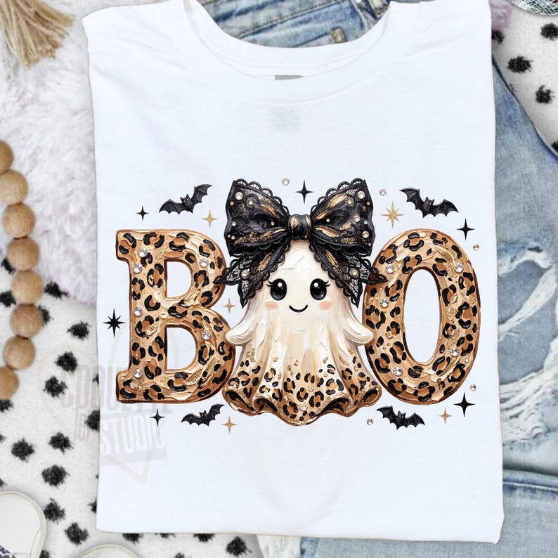 May include: A white t-shirt featuring a graphic print of the word BOO. The letters B and O are filled with a brown and black leopard print pattern. A central ghost character wears a large black lace bow and has a leopard print skirt.