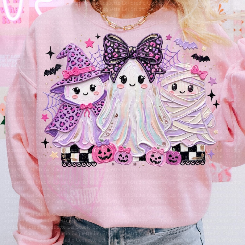 May include: A light pink sweatshirt featuring a graphic of a witch, a ghost, and a mummy. The witch wears a purple leopard print hat and cape. The ghost has a large purple leopard print bow. The mummy is wrapped in bandages. Small pumpkins are below.