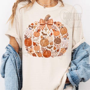 May include: A cream-colored t-shirt featuring a large central graphic of various pumpkins, autumn leaves, small ghosts, and a prominent orange and white checkered bow. The design incorporates leopard print patterns, stars, and floral elements.
