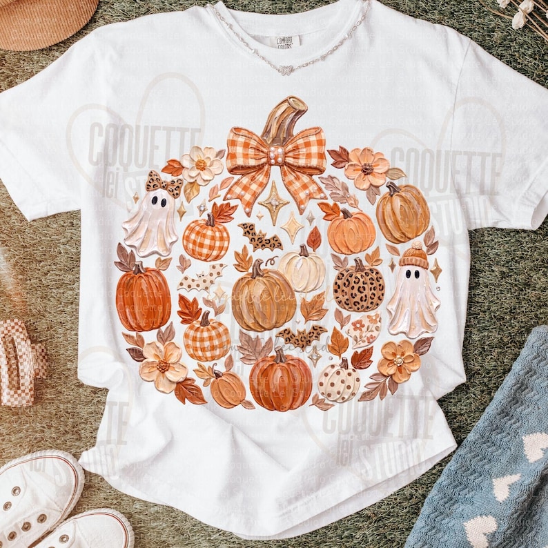 May include: A white short-sleeve t-shirt featuring a large central graphic of a pumpkin shape composed of various autumn elements including orange pumpkins, white ghosts, brown leaves, flowers, bats, and a large orange and white checkered bow.