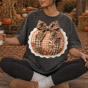 May include: Dark gray t-shirt featuring a pumpkin applique with a leopard print bow and patchwork design. The pumpkin has a white lace trim. The shirt is part of a fall-themed collection.