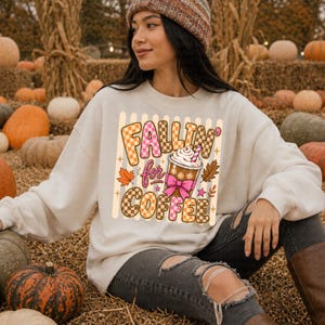 May include: A cream-coloured sweatshirt featuring the text "Fallin' for Coffee" in a checkered design. The graphic includes a coffee cup with whipped cream, a pink bow, and autumn leaves. The person is wearing a brown beanie and ripped jeans.
