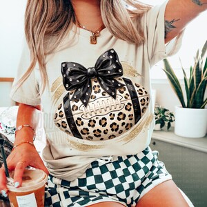 May include: A cream-colored t-shirt featuring a graphic of a football with a leopard print pattern and a large black bow with rhinestone accents. The design includes gold brushstroke details. The shirt is paired with green and white checkered shorts.