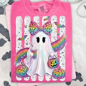 May include: A bright pink t-shirt featuring a central graphic of a white ghost wearing a large, colorful leopard-print bow. The ghost holds a patterned coffee cup and a jack-o'-lantern bucket, surrounded by rainbow wings, bats, and pumpkins.