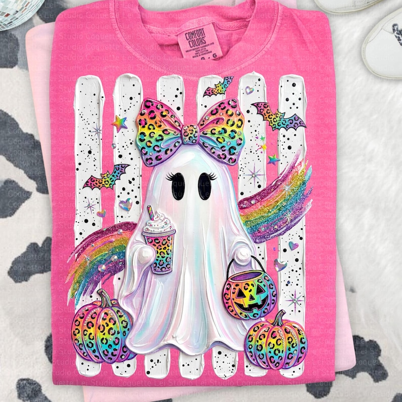 May include: A bright pink t-shirt featuring a central graphic of a white ghost wearing a large, colorful leopard-print bow. The ghost holds a patterned coffee cup and a jack-o'-lantern bucket, surrounded by rainbow wings, bats, and pumpkins.