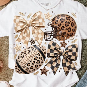 May include: A white t-shirt featuring a graphic print of a tan and white checkered bow, a brown leopard print football helmet, a tan and black leopard print football, a black and tan checkered bow, and several small leopard and checkered stars.