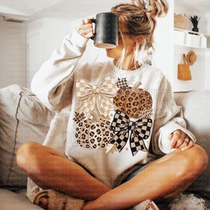 May include: A cream-colored long-sleeve sweatshirt featuring a graphic print of two pumpkins with leopard print patterns, accompanied by two large bows with checkered patterns, and small star and leaf accents, worn with a black mug.