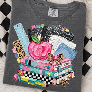 May include: Gray Comfort Colors t-shirt with a colorful school-themed design. The design includes a stack of books, an apple, a ruler, and notebooks. The apple is pink, and the books are various colors.