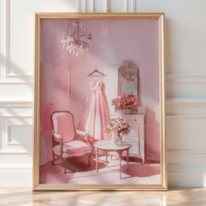 Puede incluir: Una impresión artística enmarcada representa una habitación rosa con una lámpara de araña, un vestido y una silla. Un vestido rosa cuelga de una percha de madera. Un sillón rosa y una pequeña mesa redonda están en primer plano. Una cómoda blanca y un espejo están en el fondo.