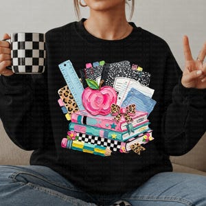 May include: Black sweatshirt featuring a colorful school-themed graphic. The design includes a pink apple, books, pencils, a ruler, and a leopard print bow. A black and white checkered mug is held in the left hand.