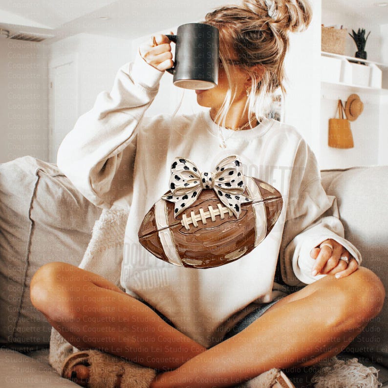 May include: A cream-colored long-sleeve sweatshirt featuring a large graphic of a brown American football adorned with a white and black spotted bow. The design is centered on the chest, showcasing a textured, painted appearance of the football.