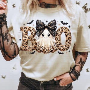 May include: A cream-colored t-shirt featuring a graphic of a ghost with a black lace bow, surrounded by the text BOO in a leopard print pattern. The design includes small black bats and star accents, with the ghost also featuring leopard print details.