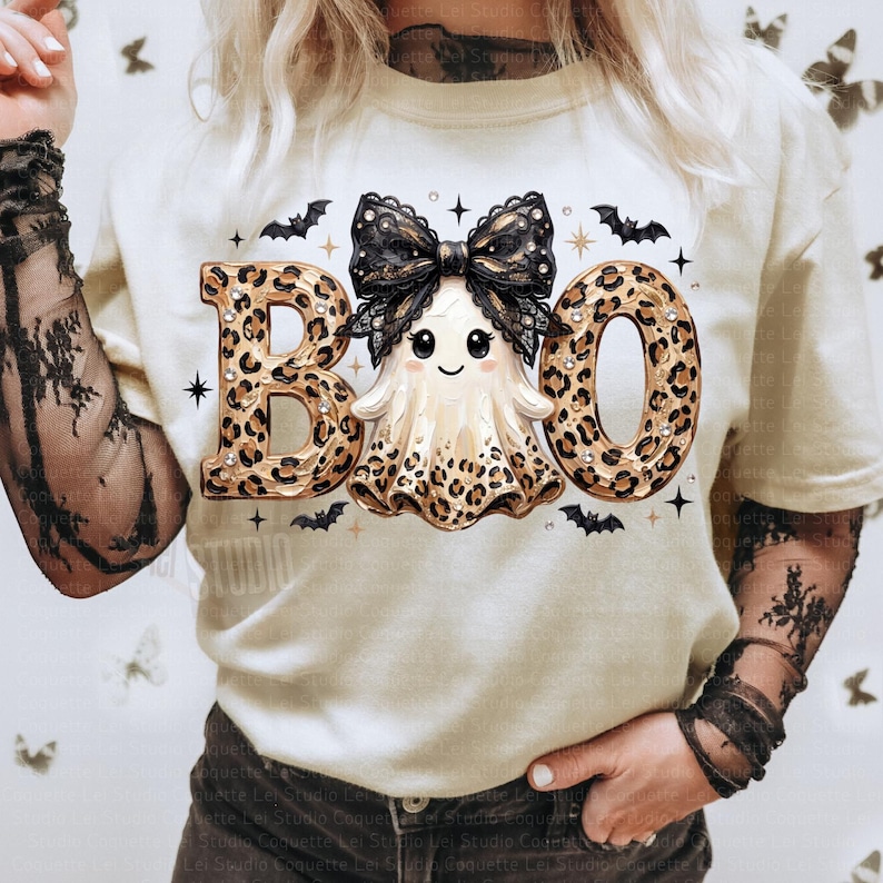May include: A cream-colored t-shirt featuring a graphic of a ghost with a black lace bow, surrounded by the text BOO in a leopard print pattern. The design includes small black bats and star accents, with the ghost also featuring leopard print details.