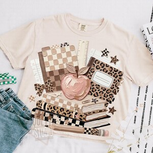 May include: Cream-colored t-shirt featuring a stack of books, an apple, and school supplies. The books have checkered, leopard, and solid patterns. A ruler, stars, and a bow are also included in the design. The text "JULY STUDIO" is visible.