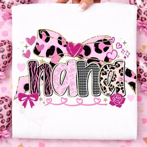 K&ouml;nnte beinhalten: Wei&szlig;es T-Shirt mit dem Wort "NANA" in verschiedenen Mustern: Zebra, floral und Leopardenmuster. Das Design umfasst eine rosa Schleife, Herzen und goldene Akzente. Eine Rose und weitere Herzen vervollst&auml;ndigen das Design.
