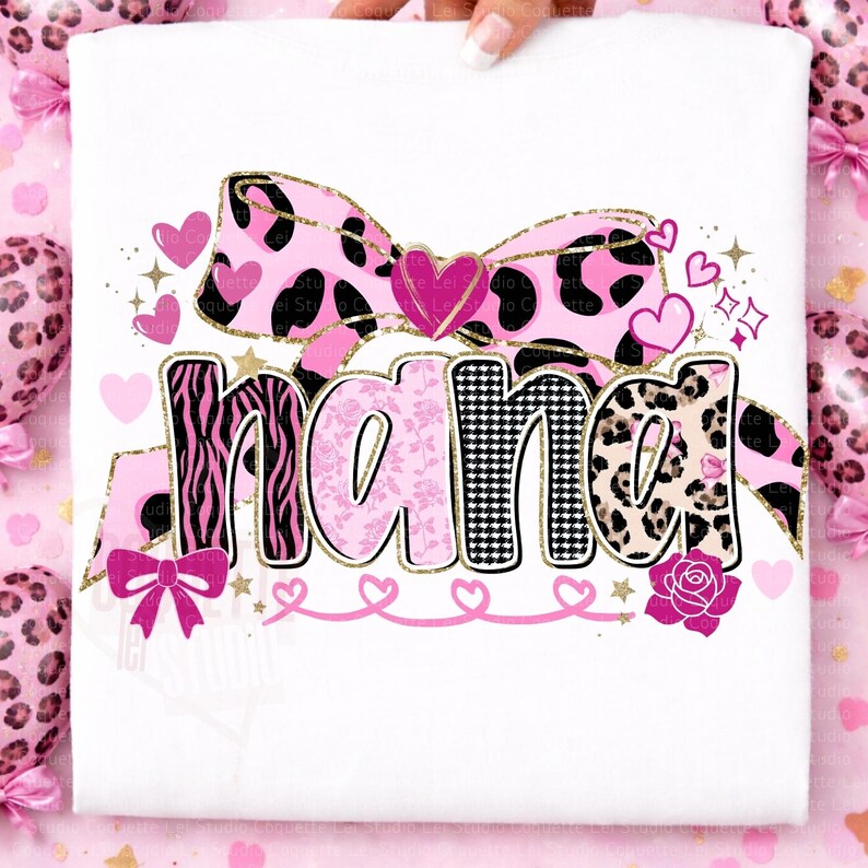 K&ouml;nnte beinhalten: Wei&szlig;es T-Shirt mit dem Wort "NANA" in verschiedenen Mustern: Zebra, floral und Leopardenmuster. Das Design umfasst eine rosa Schleife, Herzen und goldene Akzente. Eine Rose und weitere Herzen vervollst&auml;ndigen das Design.