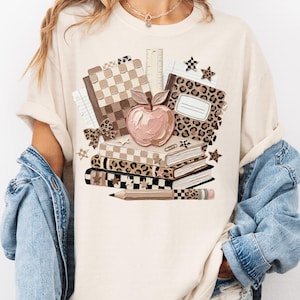 May include: Beige t-shirt featuring a stack of books, an apple, and school supplies. The books have leopard, checkered, and solid patterns. A pencil and ruler are also visible. The design is in shades of brown, beige, and pink.