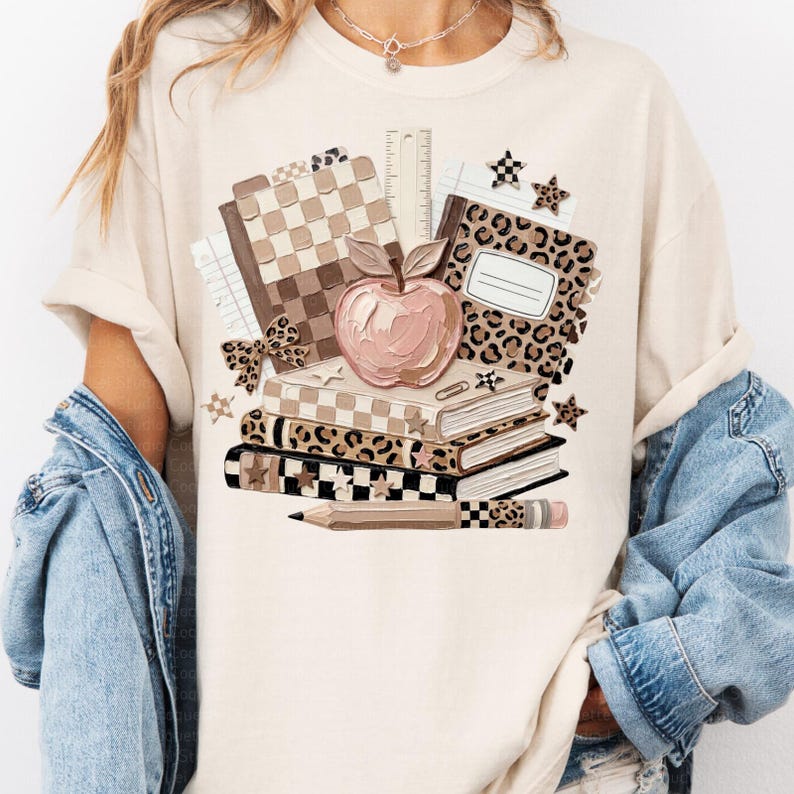 May include: Beige t-shirt featuring a stack of books, an apple, and school supplies. The books have leopard, checkered, and solid patterns. A pencil and ruler are also visible. The design is in shades of brown, beige, and pink.