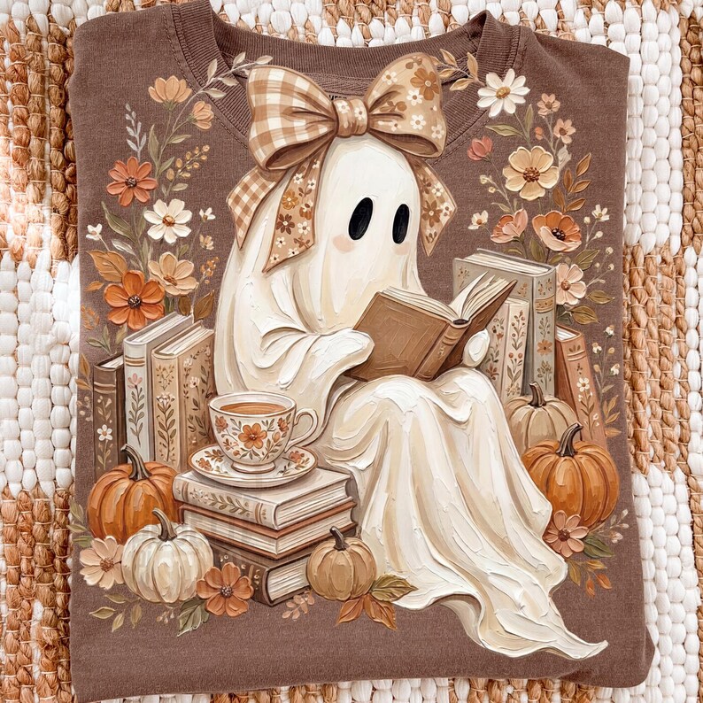 Puede incluir: Una sudadera marr&oacute;n con el gr&aacute;fico de un fantasma blanco con un lazo de cuadros beige, leyendo un libro abierto. El fantasma est&aacute; rodeado de pilas de libros, calabazas naranjas y blancas, una taza de t&eacute; floral y flores de tonos oto&ntilde;ales.